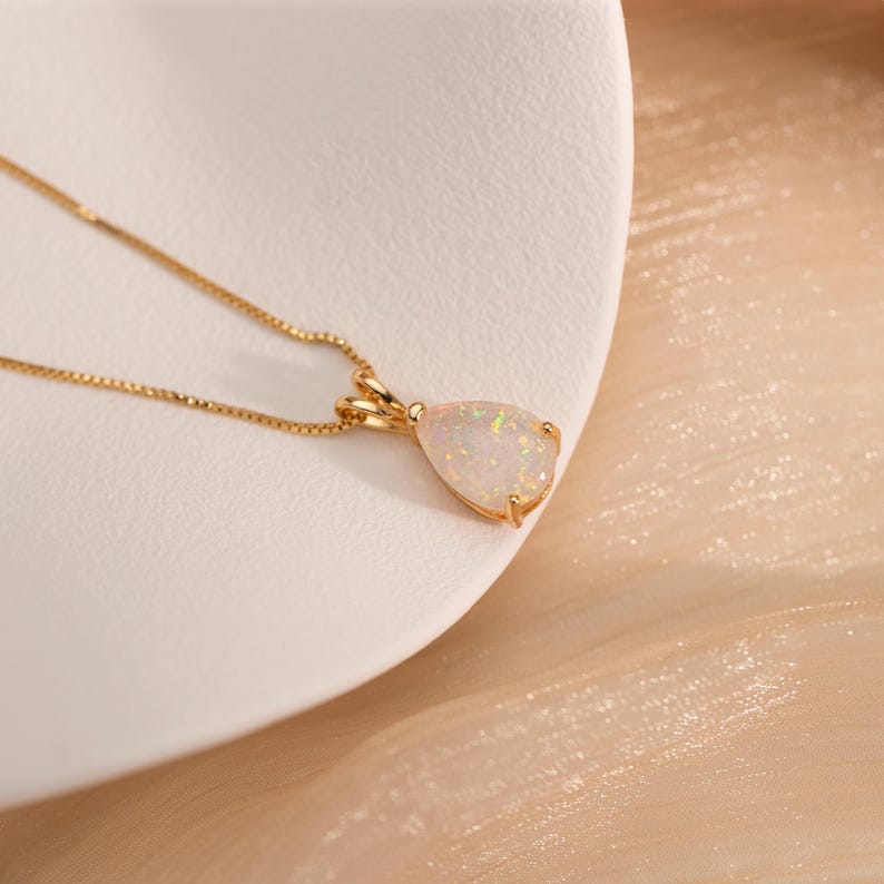 May include: A gold necklace with a pear-shaped opal pendant. The opal has a milky white base with flashes of color. The chain is a delicate gold link. The pendant is set in a gold bezel.