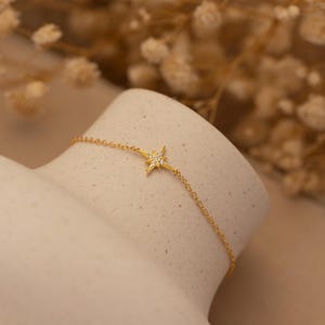 May include: A delicate gold-colored bracelet featuring a starburst pendant with small, sparkling gemstones. The bracelet is displayed on a neutral-toned surface, showcasing its fine details and elegant design. This piece of jewelry is perfect for adding a touch of sparkle.