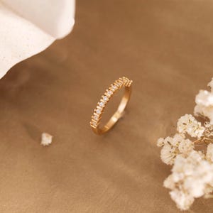 Puede incluir: Un anillo dorado con una fila de pequeñas piedras preciosas transparentes. El anillo se muestra sobre un fondo marrón, con flores blancas y un objeto blanco en el fondo. Una delicada pieza de joyería.