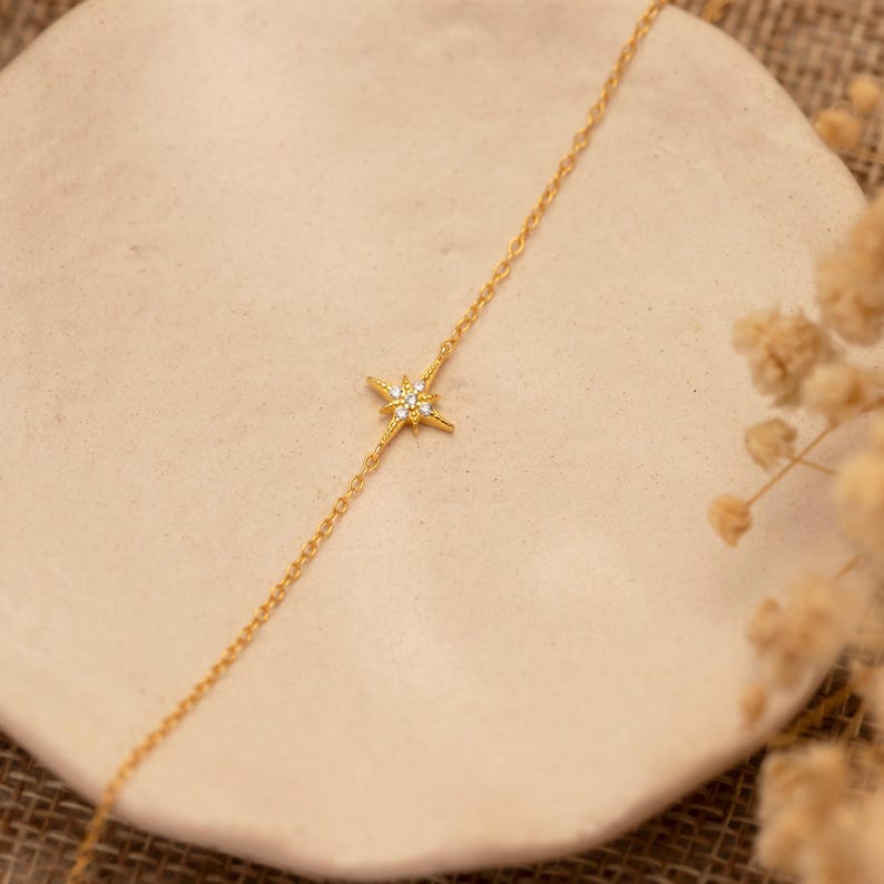 May include: A delicate gold-colored necklace featuring a starburst pendant with small, sparkling gemstones. The necklace is displayed on a neutral-toned surface, with dried floral accents in the background. The chain is fine and elegant.