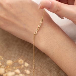 Pode incluir: Uma pulseira dourada com um design de ondas, adornada com pequenas pedras brilhantes. A pulseira tem uma corrente delicada e um motivo de três ondas. A pulseira está sendo segurada por uma pessoa.