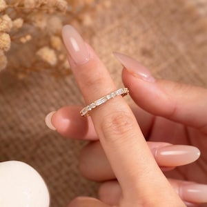 Puede incluir: Un anillo de oro con una banda de piedras claras talladas en baguette y redondas alternas. El anillo se muestra en un dedo, con un fondo borroso. El anillo es una joya.