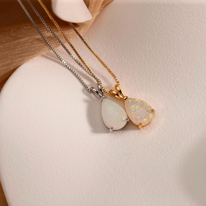 May include: Two necklaces with teardrop-shaped opal pendants. One pendant is set in silver, the other in gold. The opals display a play of color, with flashes of white, yellow, and orange. The chains are delicate.