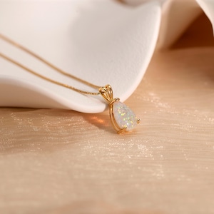 May include: A gold-colored necklace with a pear-shaped pendant. The pendant features a white opal stone with iridescent flecks. The necklace is displayed on a textured, light-colored surface.
