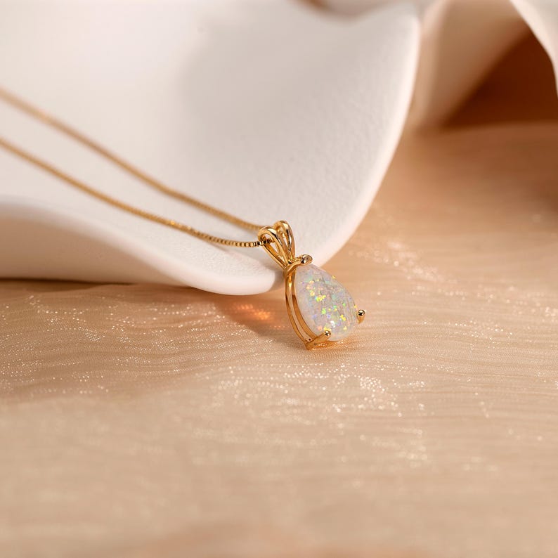 May include: A gold-colored necklace with a pear-shaped pendant. The pendant features a white opal stone with iridescent flecks. The necklace is displayed on a textured, light-colored surface.