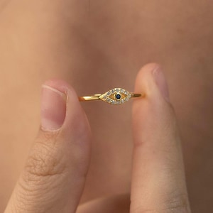 May include: A gold ring with an eye motif. The eye has a small blue centre and is encircled by tiny, clear stones. The ring is held between two fingers, highlighting its delicate design. The ring is a piece of jewellery.