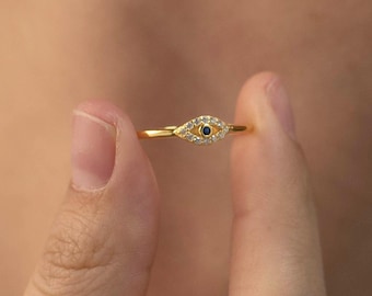 May include: A gold-colored ring featuring an eye design. The eye is detailed with a small blue center and surrounded by small, clear stones. The ring is held between two fingers, showcasing its delicate design.