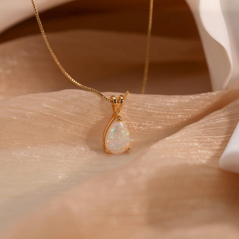 May include: A gold-colored necklace with a pear-shaped pendant. The pendant features a white opal stone with iridescent flashes of color. The necklace is displayed on a peach-colored fabric.