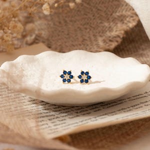 May include: A pair of gold-toned flower stud earrings. Each earring features six dark blue petals surrounding a central clear stone. The earrings are displayed on a white, textured, oval-shaped dish, resting on a piece of paper with text.