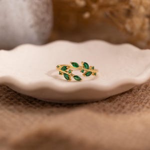 May include: A gold-colored ring featuring a leaf design with green gemstone accents. The ring is open, with the leaf design extending around the finger. The ring is set on a white, scalloped-edge plate.
