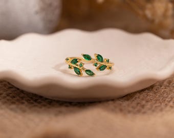May include: A gold-colored ring featuring a leaf design with green gemstone accents. The ring is open, with the leaf design extending around the finger. The ring is set on a white, scalloped-edge plate.