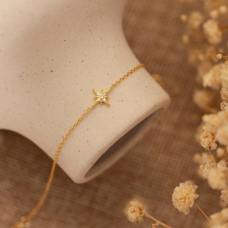 May include: A delicate gold bracelet featuring a starburst pendant with small, sparkling accents. The bracelet is displayed on a neutral-toned surface, with soft, blurred floral elements in the background. The chain is fine and elegant, suitable for everyday wear.