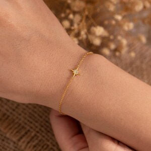 May include: A delicate gold-colored bracelet featuring a starburst charm with small, sparkling accents. The bracelet is a fine chain, and the starburst is centered on the wrist. The jewelry is displayed on a person's wrist.