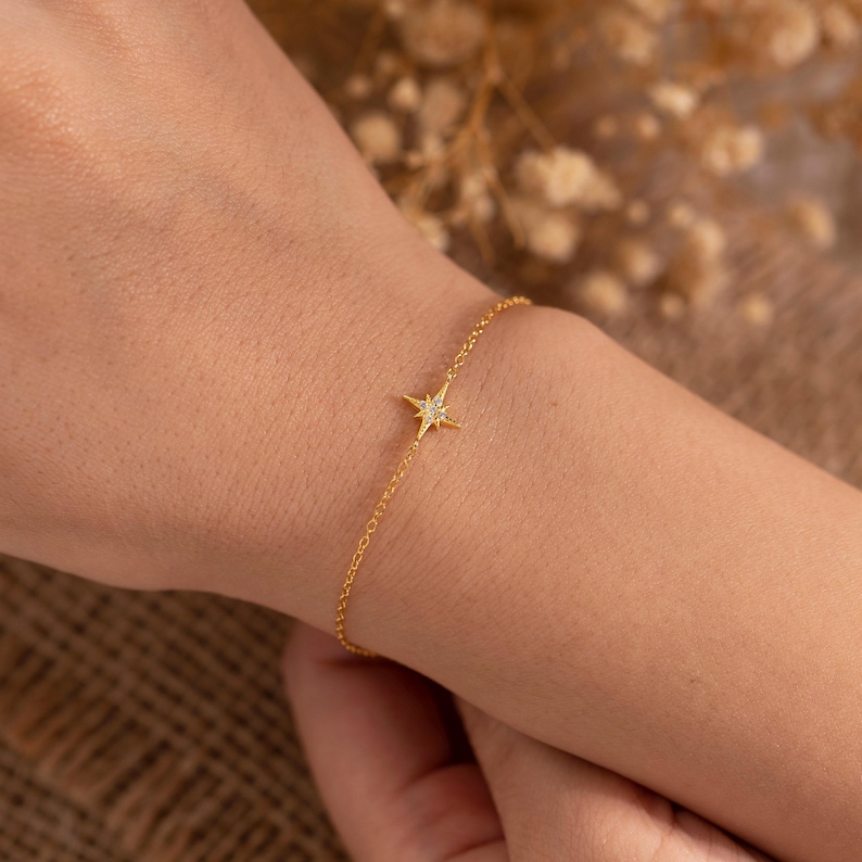 May include: A delicate gold-colored bracelet featuring a starburst charm with small, sparkling accents. The bracelet is a fine chain, and the starburst is centered on the wrist. The jewelry is displayed on a person's wrist.
