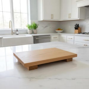 Puede incluir: Una tabla de servir de madera clara con forma rectangular, elevada sobre cuatro pequeños pies cuadrados. La veta de la madera es visible. La tabla está colocada sobre una encimera blanca en una cocina.
