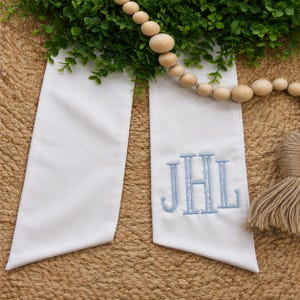 May include: A white fabric sash featuring a light blue embroidered monogram with the letters JHL. The sash has two long, flat ends with angled tips and is displayed alongside a string of natural wooden beads and a tan jute tassel.