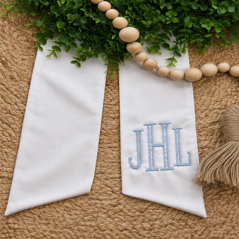 May include: A white fabric sash featuring a light blue embroidered monogram with the letters JHL. The sash has two long, flat ends with angled tips and is displayed alongside a string of natural wooden beads and a tan jute tassel.