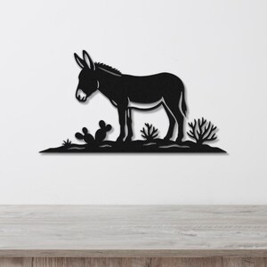 Black metal wall art silhouette of a burro standing in a desert scene with cacti, mounted on a white wall above a wooden surface.