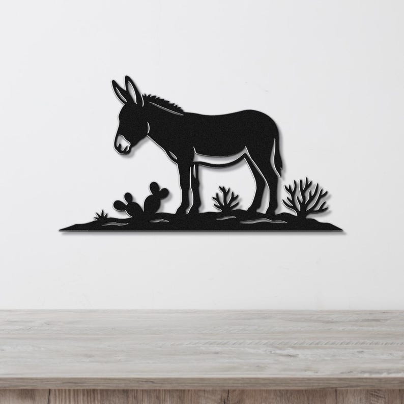 Black metal wall art silhouette of a burro standing in a desert scene with cacti, mounted on a white wall above a wooden surface.
