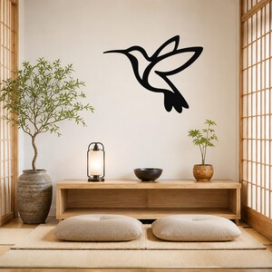 Black metal hummingbird wall art with a silhouette cutout design, mounted on a wall above a wooden console in a minimalist room.