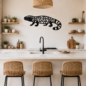 Black metal Gila monster wall art with intricate cutout patterns, mounted on a kitchen wall above a counter with wicker stools.