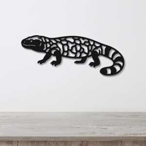 Black metal wall art of a Gila monster with intricate cutout patterns, mounted on a white wall above a wooden surface.