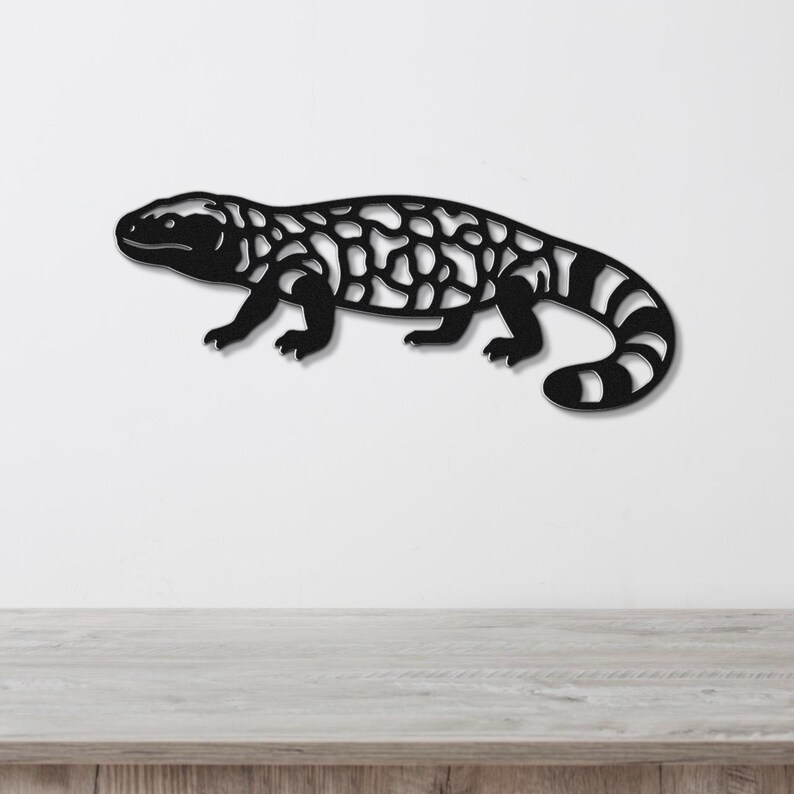 Black metal wall art of a Gila monster with intricate cutout patterns, mounted on a white wall above a wooden surface.