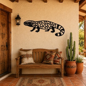 Black metal Gila monster wall art with intricate internal cutouts, mounted on a stucco wall above a wooden bench in a rustic patio setting.