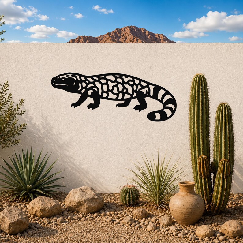 Black metal Gila monster wall art with intricate cutout patterns, mounted on a white outdoor wall above a desert garden with cacti and rocks.