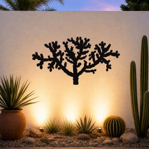 Black metal Cholla cactus wall art with branching cutout silhouette, mounted on a light-colored outdoor wall above a desert garden with potted succulents and ground lights.