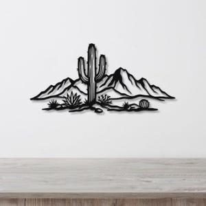 Black metal wall art silhouette of a saguaro cactus and desert landscape with mountains and smaller cacti, mounted on a white wall above a light wood surface.