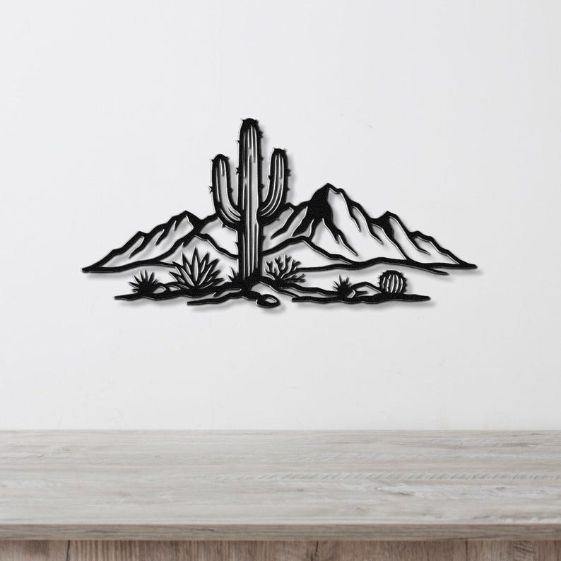 Black metal wall art silhouette of a saguaro cactus and desert landscape with mountains and smaller cacti, mounted on a white wall above a light wood surface.