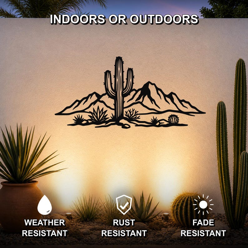 Black metal wall art featuring a desert landscape silhouette with a saguaro cactus and mountains, mounted on an outdoor wall above a gravel garden with potted cacti.