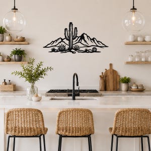 Black metal wall art featuring a saguaro cactus and mountain silhouette, mounted above a kitchen counter with open shelving and pendant lights.