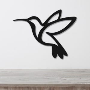 Black metal hummingbird wall art with a silhouette cutout design, mounted on a white wall above a light wood surface.
