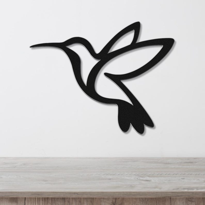 Black metal hummingbird wall art with a silhouette cutout design, mounted on a white wall above a light wood surface.