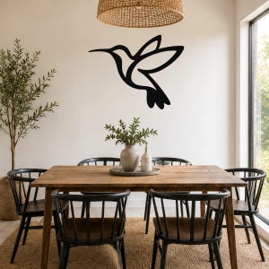 Black metal hummingbird wall art with motion-line cutouts, mounted on a white wall above a wooden dining table in a bright dining room.