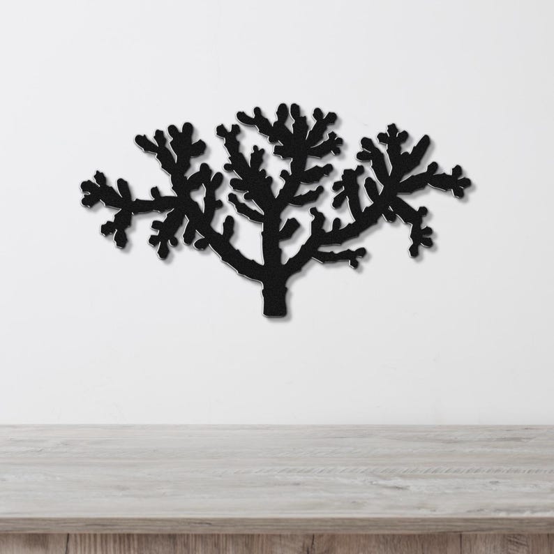 Black metal wall art cutout of a cholla cactus with branching arms, mounted on a white wall above a light wood surface.