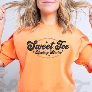 May include: Orange t-shirt with the text "Sweet Tee Mockup Studio" in a black, stylized font. The shirt is short-sleeved and has a round neckline. The model is wearing denim shorts.