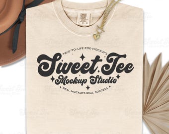 May include: Cream-colored sweatshirt with the text "Sweet Tee Mockup Studio" in a retro font. A gold necklace with a heart pendant rests on the shirt. A brown hat and dried palm leaves are in the background.