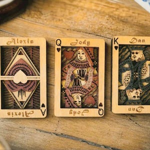 May include: Three rectangular wooden playing cards featuring laser-cut designs. The left card is an Ace of Hearts labeled Alexis, the center is a Queen of Hearts labeled Jody, and the right is a King of Hearts labeled Dan, all with intricate details.