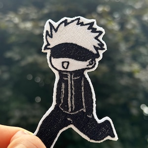 May include: Embroidered patch featuring a cartoon character in a black outfit with white hair and a blindfold. The character is depicted in a running pose, with a white border around the design. The patch is held against a blurred green background.
