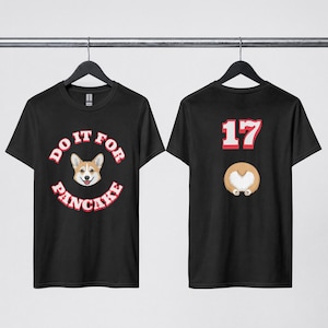 May include: A black t-shirt featuring a front graphic of a corgi face surrounded by the text DO IT FOR PANCAKE in white and red. The back of the shirt displays the number 17 in red and white above a graphic of a corgi rear end with a white heart.