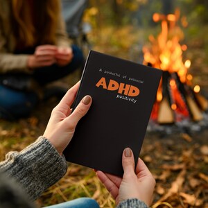 May include: A black notebook with the words "A pocketful of potential ADHD positivity" in orange and white. The notebook is held in front of a blurred background of a campfire and a person.