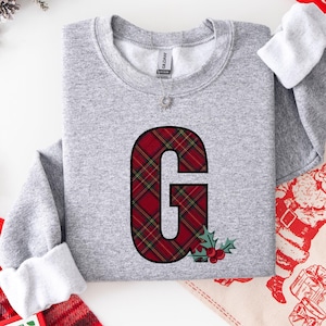May include: A gray sweatshirt featuring a large letter "G" in a red and green plaid pattern. The letter is outlined in black and adorned with holly and berries. The sweatshirt has a crew neck and long sleeves.