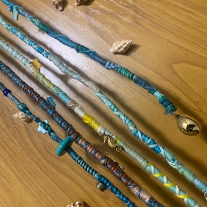 May include: Close-up of several handmade dreadlock wraps in shades of blue, teal, and brown, adorned with small charms and seashells. The wraps are arranged diagonally on a wooden surface, showcasing their intricate designs.