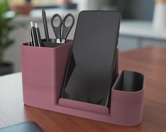 May include: A mauve desk organizer with compartments for pens, pencils, scissors, and a smartphone. The organizer is made of a solid material and sits on a wooden surface.
