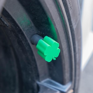 May include: A bright green, four-leaf clover valve stem cap is attached to a black tire rim. The clover-shaped cap adds a decorative touch to the vehicle's wheel. The tire is black and the background is blurred.