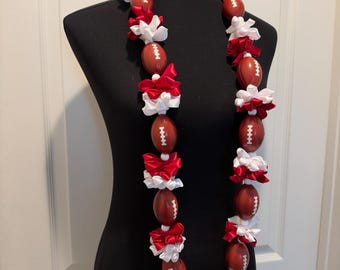 May include: A decorative lei featuring alternating brown football-shaped ornaments with white laces and clusters of red and white satin ribbon bows. The footballs are arranged in a continuous loop, interspersed with small white beads and ribbons.