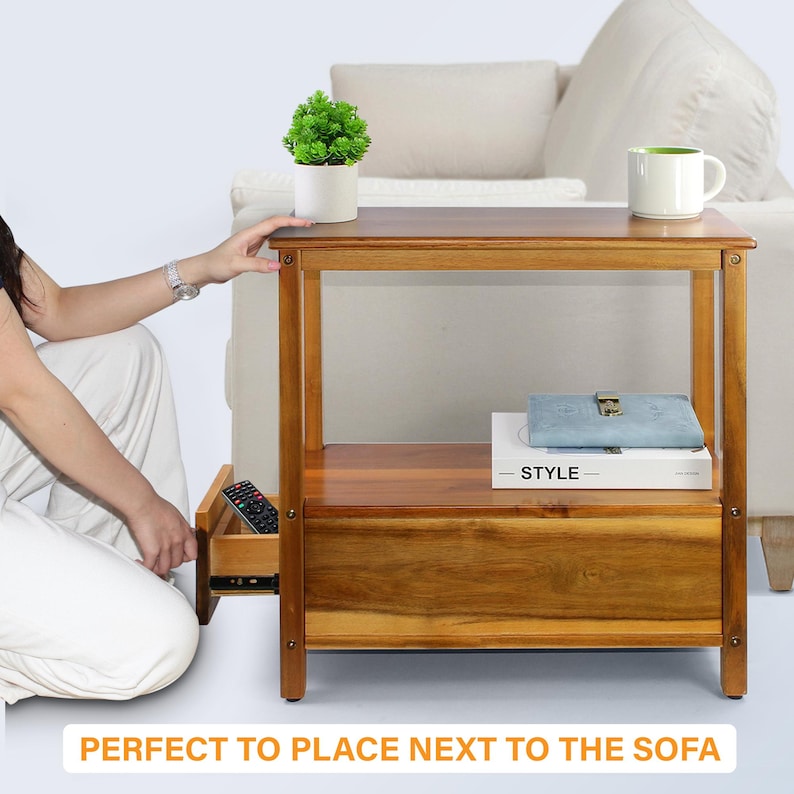 May include: A wooden side table with a drawer, a shelf, and a top surface. The table is made of a warm-toned wood and has a rectangular shape. A small potted plant and a white mug sit on top. The drawer is open, revealing a remote control.